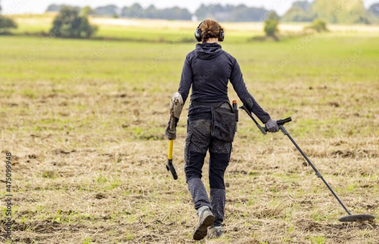 A person wearing headphones and dark clothing walks through a field with a metal detector in one hand and a shovel in the other, demonstrating advanced metal detecting techniques for gold prospectors.
