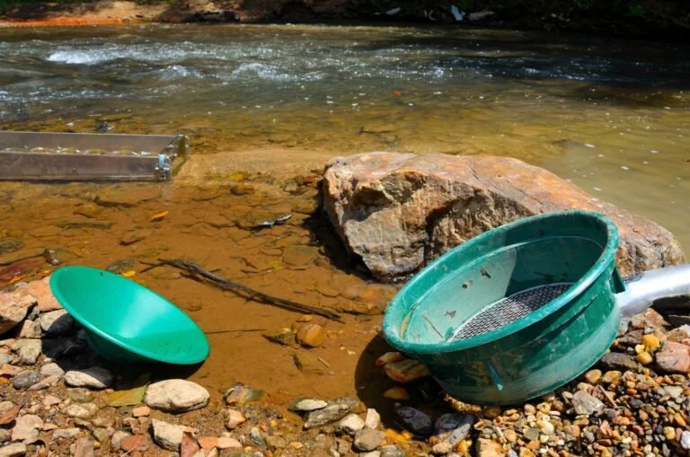 Two green gold pans and a river sluice box, known as some of the best tools for gold panning, rest on the rocky riverbank, suggesting an active search for gold near the water.