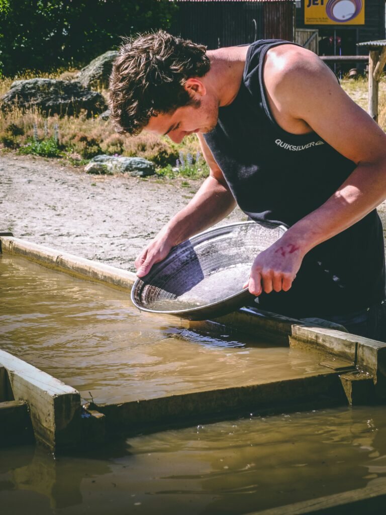 A man pans for gold in a muddy trough, holding a metal pan over water while standing outdoors on a sunny day—remembering common mistakes to avoid in your early gold panning adventures.