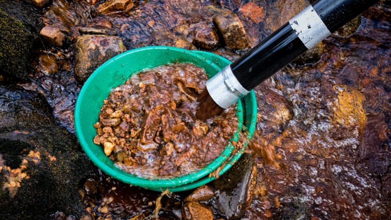 A green pan filled with rocks and gravel is being washed with a stream of water from a black hose in a shallow, rocky stream—perfect for those interested in customizing gold panning kits for their next adventure.