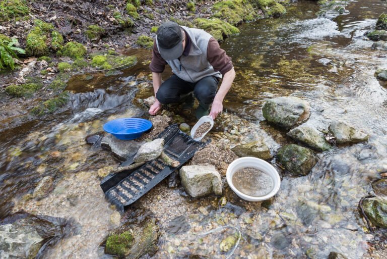 A person kneels by a shallow stream, using pans, a sluice box, and techniques inspired by DIY Gold Processing Plants to search for gold among rocks and gravel.