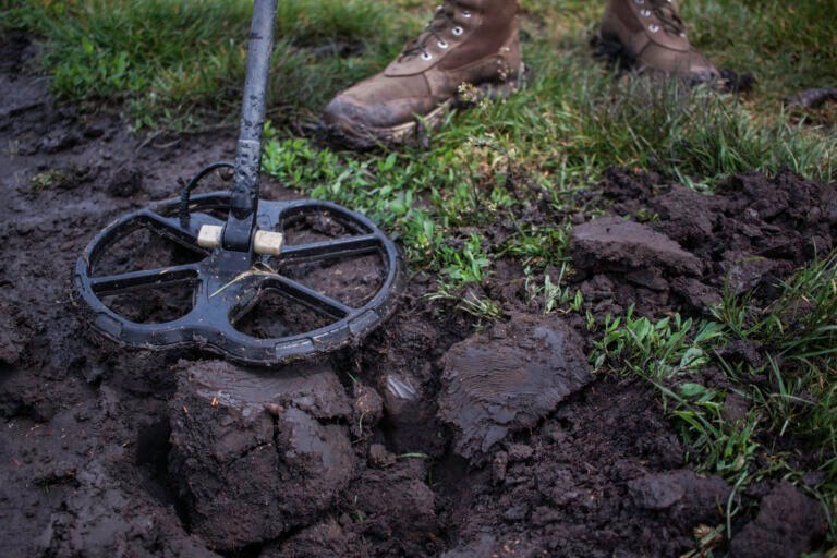 A person searching for detecting gold nuggets uses a metal detector to scan muddy ground, with patches of grass and boots visible in the scene.