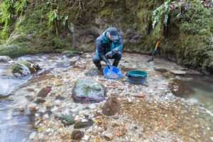 A person kneels by a shallow stream, panning for gold with a blue pan. Nearby, a green bucket, a shovel, and different types of sifting screens are placed on rocky ground.