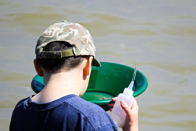 A child wearing a camouflage cap holds a green pan and a plastic bottle while standing near murky water, engaging in Purposeful Gold Panning Education for Kids.