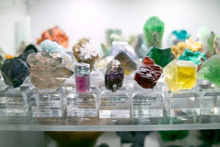 Various colorful mineral and crystal specimens, often uncovered during Precious Metals Exploration, are displayed on clear stands, each labeled with their name and origin.