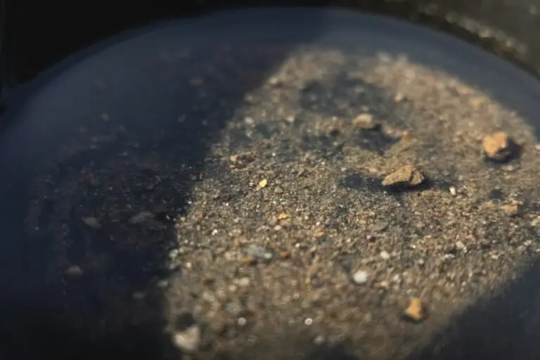 Close-up of wet sand and small rocks, reminiscent of gemstones in gold panning, partially submerged in water with a shadow cast across the surface.