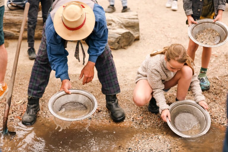 Two people, one adult and one child, are panning for gold in a shallow stream using metal pans, beginning their own Adventure Tales. Other people are visible in the background.