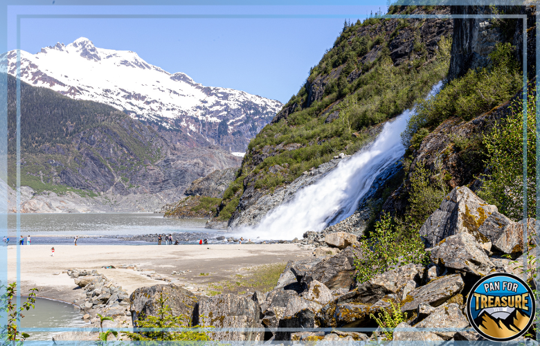 A waterfall flows down a rocky, tree-covered mountain slope near a lake, where people stroll along the sandy shore and discuss Gold Panning Laws in Alaska against a backdrop of snow-capped peaks.