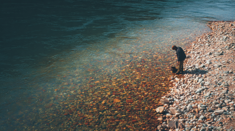 Gold Panning Safety A person stands on a rocky riverbank, looking down and holding a bucket near the edge of clear, shallow water—practicing gold panning safety while searching for treasures.