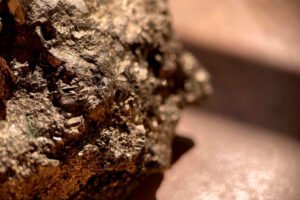 Close-up view of a rough, metallic mineral rock, possibly pyrite, with a textured and reflective surface on a smooth, light brown background—suggestive of materials used in gold refining.