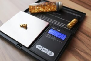 A small gold nugget is being weighed on a digital scale using gold testing equipment, with two vials of gold flakes placed nearby on a wooden surface.