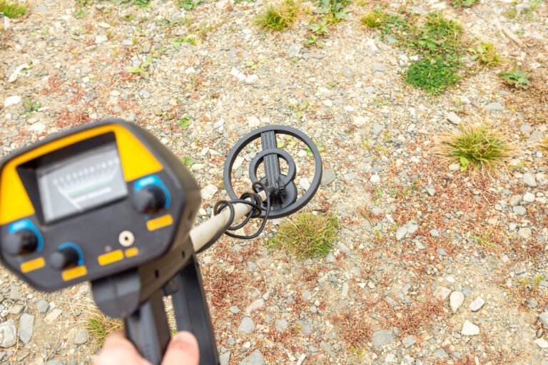 A handheld metal detector is being used to scan rocky ground with sparse grass, demonstrating how to find gold with a metal detector in challenging terrain.