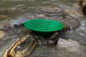 A green plastic gold panning pan sits on rocks in a shallow, flowing stream, demonstrating effective gold pan usage for prospectors seeking treasure.