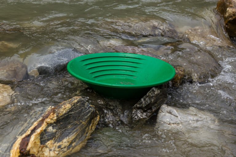 A green plastic gold panning pan sits on rocks in a shallow, flowing stream, demonstrating effective gold pan usage for prospectors seeking treasure.