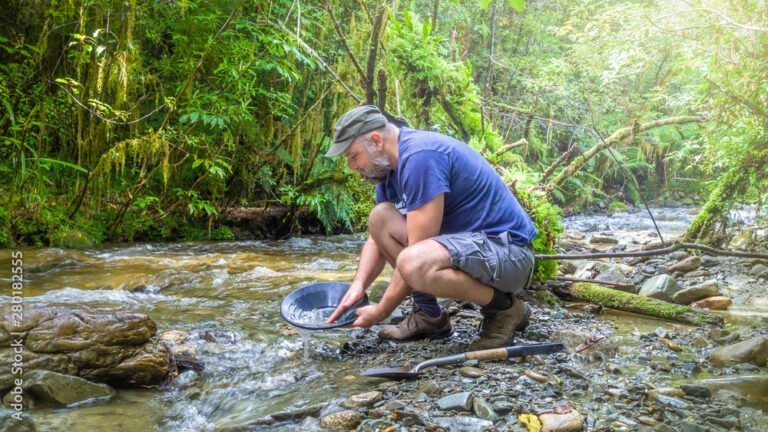 A man crouches at the edge of a rocky creek in a forest, demonstrating how to gold pan with a black pan. Tools for panning lie on the ground nearby.