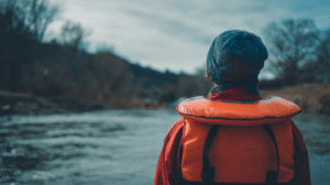 Person wearing an orange life jacket and blue beanie looks out over a calm river, practicing water safety in gold panning, with trees and hills in the background under a cloudy sky.