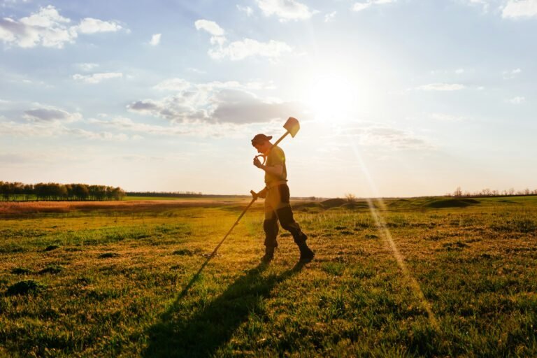 A person walks across a grassy field at sunset, carrying a shovel over their shoulder, ready for some Metal Detector Maintenance.
