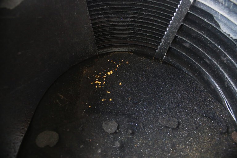 Small gold flakes and micro-gold are visible at the bottom of a black ribbed pan filled with dark sediment, commonly used in gold panning.