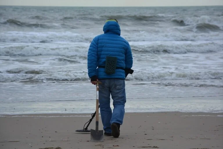 Person in a blue jacket and jeans using a metal detector and holding a shovel walks along a sandy beach near the ocean waves, searching for coins, jewelry, and other valuables you can find with a metal detector.