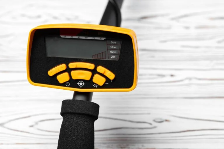 A close-up of a yellow and black metal detector control panel with digital display and buttons, designed for pinpointing gold with a detector, on a white wooden surface.