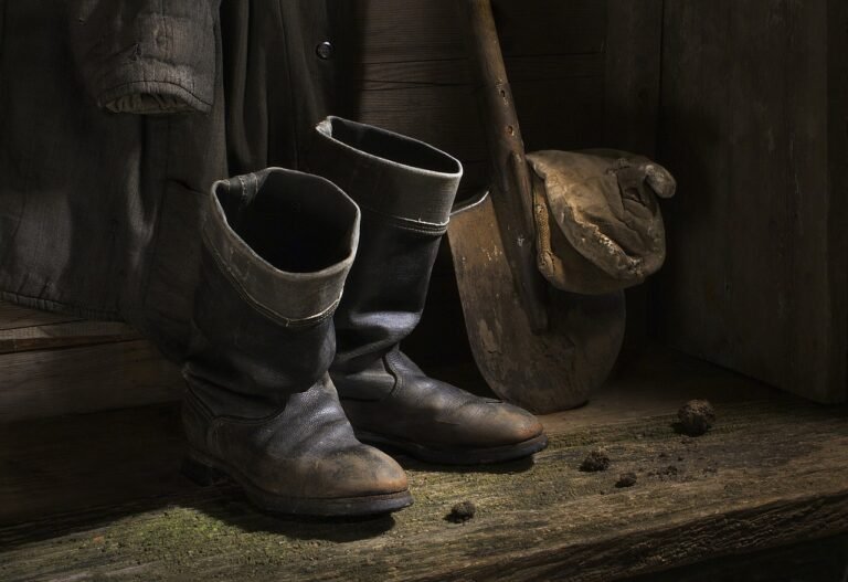 A pair of worn black boots, a coat, and a muddy shovel—remnants of Panning Tour Experiences—rest against a wooden wall on a weathered wooden floor.