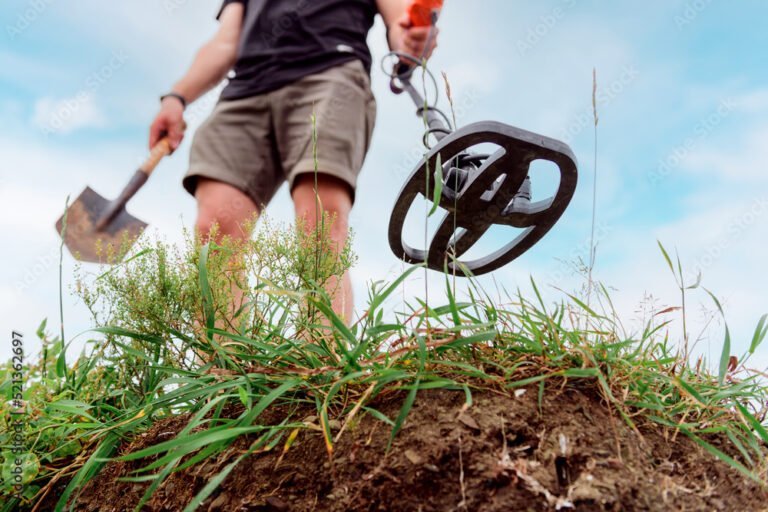 Person using a metal detector and holding a shovel, searching for objects in the soil outdoors with grass and cloudy sky visible, illustrating the real-world use of Pulse Induction vs. VLF Detectors in treasure hunting.