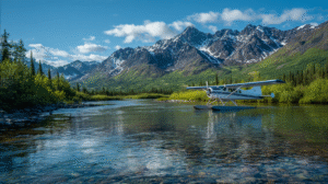 A floatplane sits on a clear, shallow river ideal for responsible gold panning, with green trees and snow-capped mountains in the background under a blue sky.