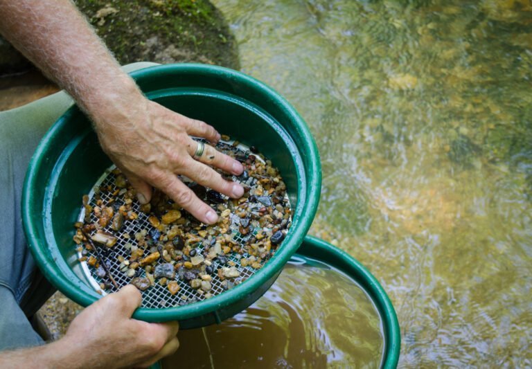 A person, practicing sifter and classifier maintenance, skillfully sifts rocks and gravel in a green pan over water—likely panning for gold or gemstones near a stream.