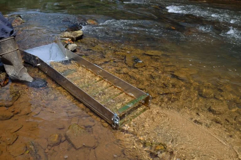 A metal gold sluice box is placed in a shallow, rocky stream with water flowing through it for gold prospecting, offering a more efficient alternative when comparing Sluice Box vs. Gold Pan techniques.