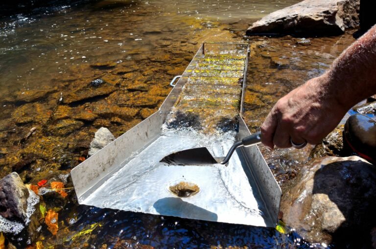 A person uses a trowel to extract material from a sluice box placed in a shallow stream, demonstrating sluicing best practices for successful gold prospecting.