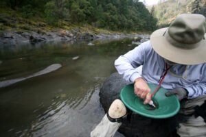 A person wearing outdoor gear uses specialized equipment for fine gold as they pan in a shallow river, surrounded by forest and rocks.