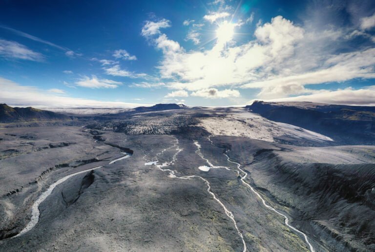 Aerial view of a volcanic landscape shaped by tectonic activity in gold formation, with winding rivers, rocky terrain, and distant mountains under a sunny, partly cloudy sky.