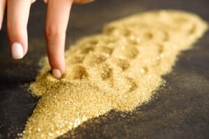 A person's hand traces patterns in a pile of golden granulated sugar scattered on a dark surface, resembling a Fine Gold Classification.