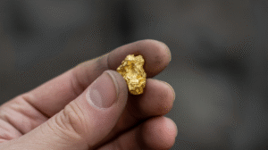 A hand holding a small, rough gold nugget between the thumb and index finger with a blurred background, showcasing the results of Nugget Prospecting Techniques.