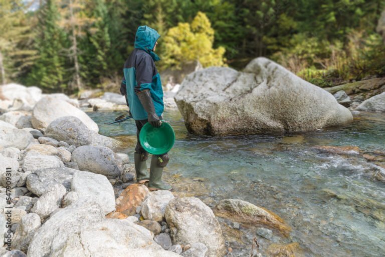A person in outdoor gear stands on rocks by a river, holding a green pan—like the ones found in Kids' Gold Panning Kits—and looking at the water, surrounded by trees and large boulders.