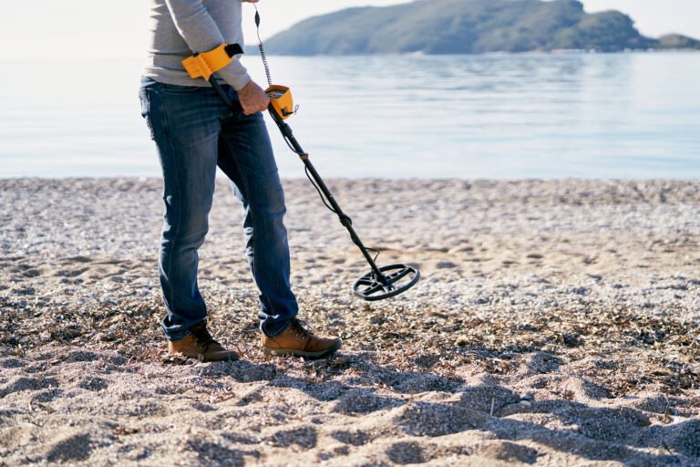 Person troubleshooting detector issues while using a metal detector on a pebbly beach near the shoreline, with water and distant hills in the background.