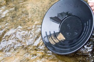 A person from a Gold Pans Selection holds a black gold pan with sediment and water over a shallow stream.