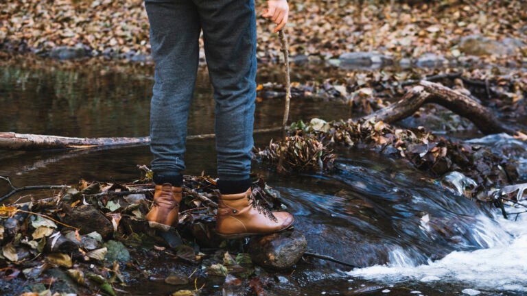 Person wearing brown boots and gray pants stands on rocks and leaves by a small creek, holding a stick, with water flowing over stones—wondering, where is gold most likely to be found in such a setting?.
