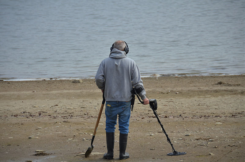 A man standing on the beach with a shovel, wondering if gold can be detected by a metal detector.