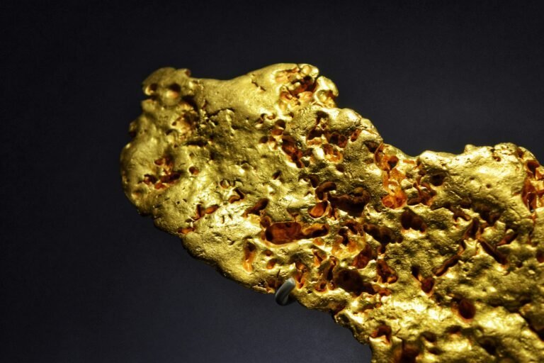 A close-up of a gold nugget with a rough, porous surface set against a dark background, revealing unique textures created through natural gold formation.